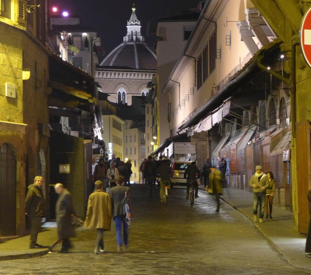 Night street-level view along the Ponte Vecchio bridge deck between the (shuttered) jewellers' shops, with the Duomo dome beyond