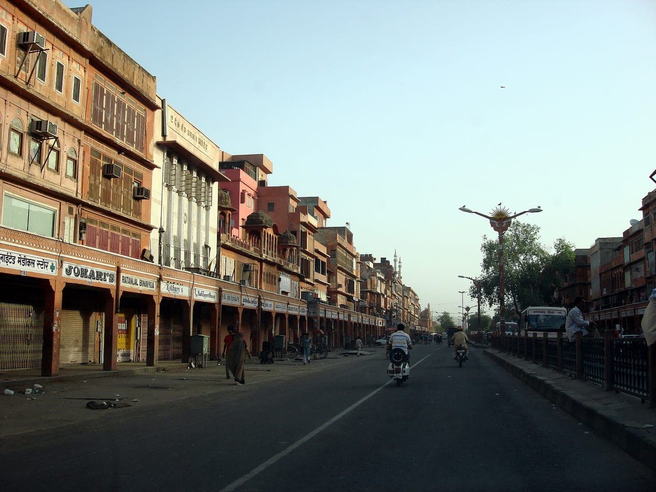Street view in Jaipur's walled Pink City — arcaded pink-façade shopfronts of the old-city jewellers' bazaar (a 'JOHARI'S' jeweller sign visible)
